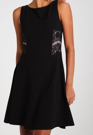 Ärmelloses schwarzes Kleid mit ausgestelltem Rock und Spitzenpaneeleinsätzen an den Seiten, getragen von einer Person, die vor einem einfarbigen Hintergrund steht.