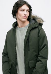Grøn hætteparka med falsk pelskant, lynlås og sidelommer, båret over en lys grå crewneck sweater. Glat, tykt stof.