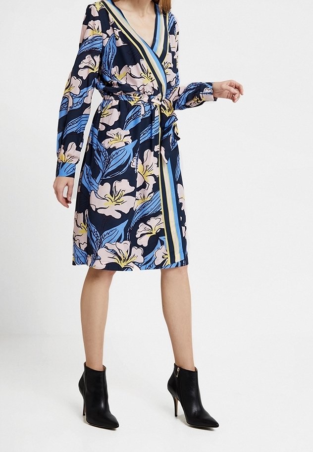 Robe portefeuille avec un motif floral bleu marine, des fleurs roses et des feuilles bleues. Manches longues, décolleté en V et taille nouée. Associée à des bottines noires à la cheville.