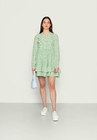 Vestido florido verde com mangas compridas, pregas e saia em camadas. Combinado com sapatos brancos e uma mala azul clara. Textura suave.