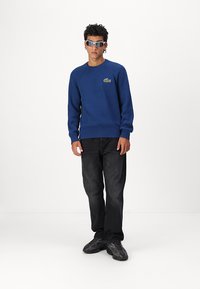 Blauer gerippter Pullover mit grünem Logo, kombiniert mit schwarzen Jeans und schwarzen Sneakers. Das Model trägt eine Sonnenbrille und steht vor einem weißen Hintergrund.