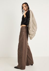 Pantalones de pierna ancha marrones con bolsillos de parche, combinados con un top corto negro y una chaqueta bomber marrón claro colgada sobre un hombro.