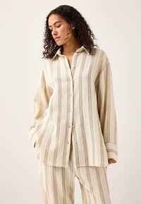 Koszula na guziki z jasnobeżowego materiału z cienkimi pionowymi paskami w kolorze szarym. Posiada kołnierzyk, długie rękawy oraz luźny krój.