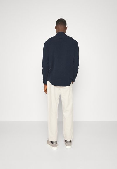 Camicia a maniche lunghe blu navy con colletto button-down, abbinata a pantaloni beige chiaro e scarpe da ginnastica grigie. Tessuto liscio, vestibilità rilassata.