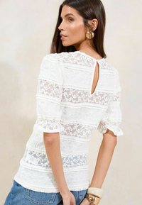 Blusa bianca in pizzo con maniche a palloncino corte, caratterizzata da un'apertura a chiave sul retro e motivi a righe orizzontali. Abbinata a jeans blu.