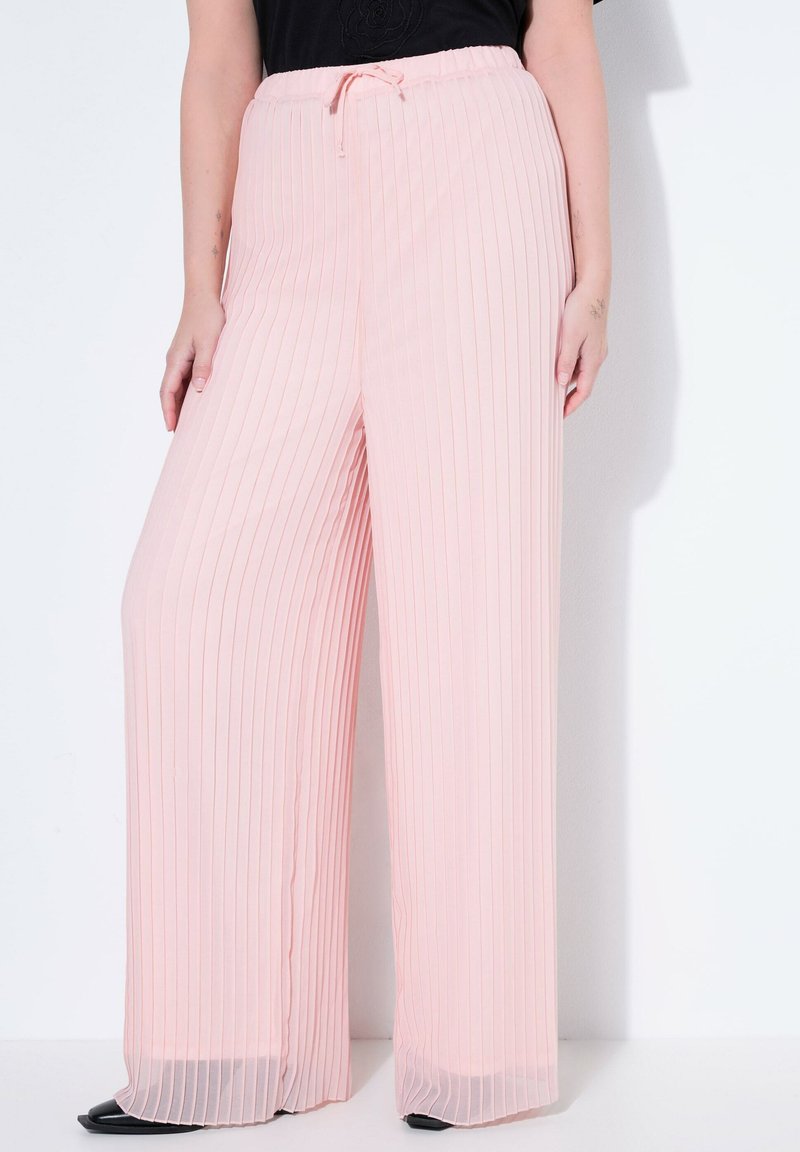 Frau trägt hochgeschnittene, hellrosa plissierte Hosen mit weitem Bein und Kordelzug in der Taille, schwarze Schuhe und ein schwarzes Oberteil.