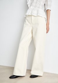 Λευκά παντελόνια σε φαρδιά γραμμή από ανθεκτικό ύφασμα με λεία υφή, σε συνδυασμό με μια ανοιχτόχρωμη, καρό μπλούζα peplum και μαύρες φλατ παντόφλες.