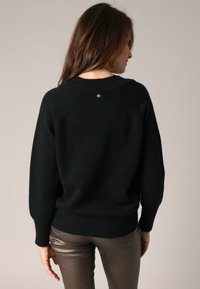 Femme aux longs cheveux bruns portant un pull noir et un pantalon brun métallique, de dos devant un fond clair et uni.