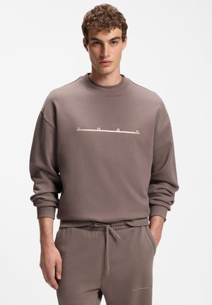 Jeune homme portant un sweat-shirt et un pantalon de jogging taupe assortis avec un logo subtil "HUGO" sur la poitrine et les mains dans les poches.