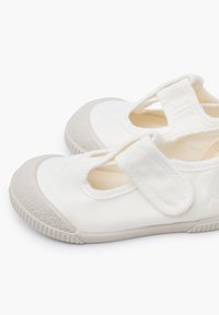 Witte canvas schoenen met een ronde neus, grijze rubberen zool en een klittenbandsluiting over de bovenkant. Voorzien van uitsparingen voor ventilatie.