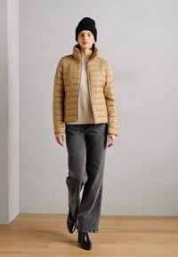 Tan pufferjakke med vandrette stikmønstre, parret med en beige top, grå vidbenede jeans og sorte ankelstøvler. Modellen bærer en sort hue.