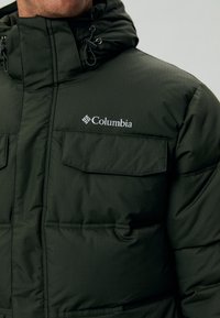Donkergroene gewatteerde jas met een ritssluiting aan de voorkant, verstelbare capuchon en twee borstzakken. Bevat het Columbia-logo aan de linkerkant. Gestructureerde stof.