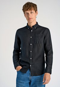 Camicia nera a maniche lunghe in tessuto di lino con colletto a punta, taschino sul petto e bottoni bianchi, abbinata a jeans in denim blu.