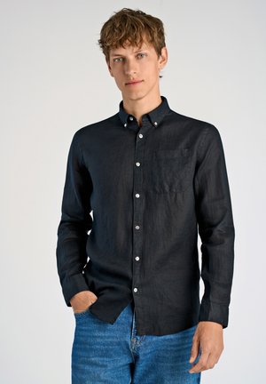 Camisa negra de manga larga en tejido de lino con cuello abotonado, bolsillo en el pecho y botones blancos, combinada con unos jeans de mezclilla azules.
