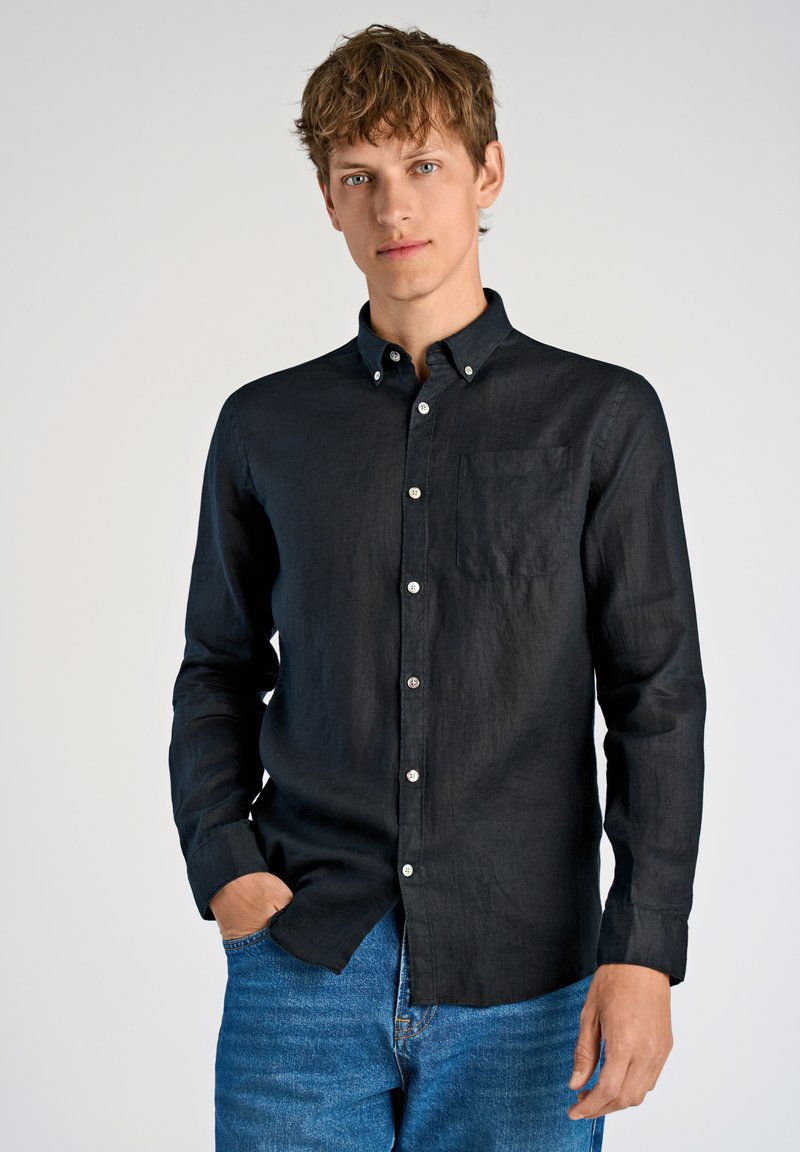 Camicia nera a maniche lunghe in tessuto di lino con colletto a punta, taschino sul petto e bottoni bianchi, abbinata a jeans in denim blu.