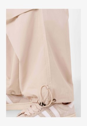 Beige Hose aus leichtem Stoff, mit einem Kordelzug am Saum, kombiniert mit beigen Sneakers, die weiße Streifen haben.