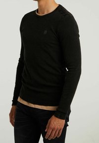 Czarna, teksturowana koszulka z długim rękawem i okrągłym dekoltem, z gładkim panelem na ramionach i małym logo na klatce piersiowej. Warstwowa, noszona na beżową podkoszulkę.