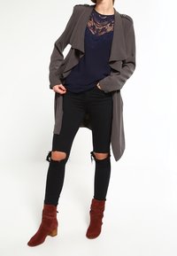 Manteau long gris porté sur un haut bleu marine avec des détails en dentelle, associé à un jean skinny noir déchiré et des bottines rouges à talon carré.