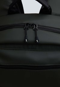 Ciemnozielony plecak o gładkiej teksturze, z dwoma czarnymi zamkami błyskawicznymi z numerowanymi uchwytami oraz dolną kieszonką na zamek z siateczkowym wykończeniem.