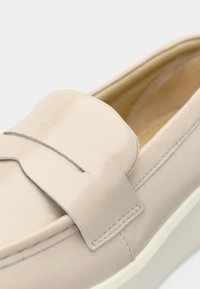 Μπεζ δερμάτινο loafer με λουρί για κέρμα και λευκή σόλα, με λεπτομερή ραφή και λείο φινίρισμα στο πάνω μέρος.