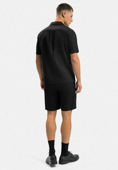 Svart kortärmad skjorta med krage, tillsammans med svarta shorts och svarta skor, visar en enkel, enhetlig design bakifrån.