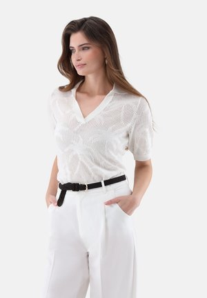 Frau mit braunen Haaren, die ein weißes, strukturiertes Kurzarm-Strickoberteil, weiße hoch taillierte Hosen und einen schwarzen geflochtenen Gürtel trägt, Hände in den Taschen.