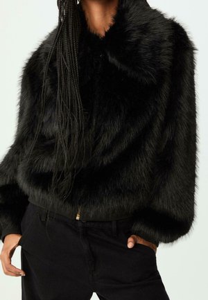 Personne avec de longues tresses portant une veste en fausse fourrure noire avec un large col et un pantalon noir, une main dans la poche.