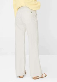 Persona che indossa pantaloni bianchi a vita alta e a gamba larga, maglione giallo e sandali bianchi, in piedi davanti a uno sfondo chiaro e uniforme.