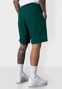 Man wearing green shorts and white sneakers, showcasing tattooed legs and a relaxed fit. The background is light and neutral.