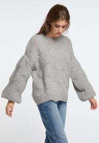 Světle šedý pletený svetr s texturovaným vlnitým vzorem, oversized rukávy a volný střih, doplněný o modré džíny.
