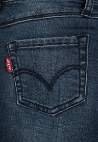 Tmavě modré džíny z denimu s zadní kapsou se specifickým vzorem V-stehu a červenou značkovou visačkou na okraji. Hladký povrch.