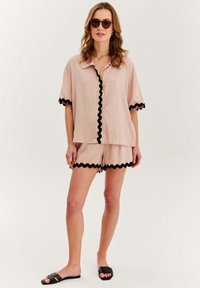 Svetloružová košeľa s krátkym rukávom a zodpovedajúce šortky s čiernym vlnitým lemom. Modelka v čiernych šľapkách a slnečných okuliaroch.