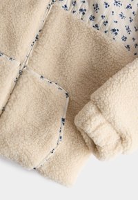 Giacca in pile di colore crema con una superficie testurizzata. Presenta una fodera floreale blu e piping a contrasto lungo le tasche e le cuciture.