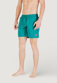Pantaloni da bagno turchesi con vita elastica, chiusura con cordoncino e una toppa con logo a sinistra. Il tessuto appare leggero e liscio.