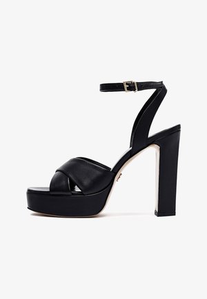 Sandalia de plataforma negra con tacón alto y grueso, con tiras cruzadas y hebilla en el tobillo, vista lateral sobre fondo blanco.