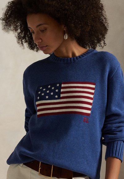 Frau mit lockigem Haar, die einen blauen Strickpullover mit amerikanischem Flaggenmuster und beige Hosen trägt, schaut seitlich nach unten.