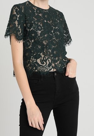 Femme portant un top court à manches courtes en dentelle florale noire sur une couche de base nude, associé à un pantalon noir taille haute, debout avec une main dans la poche.