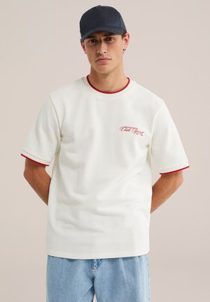 Jeune homme portant un t-shirt blanc avec un liseré rouge et du texte, un jean bleu clair et une casquette de baseball bleu foncé, debout les mains derrière le dos.