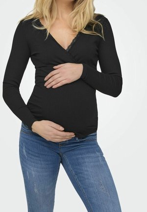 Femme enceinte portant un haut noir à manches longues et un jean bleu, tenant doucement son ventre avec les deux mains.