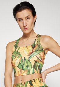 Crop top s cvjetnim uzorkom u žutoj boji s zelenim listovima i narančastim akcentima. Izrađen od rastezljivog materijala, ima zaobljeni izrez i široke naramenice.