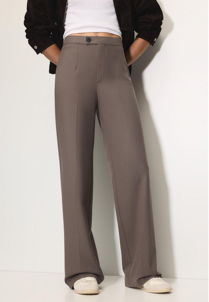 Világosbarna, bő szárú nadrágok redőkkel, gombos záródással és puha anyaggal, fehér felsővel és bézs sportcipőkkel párosítva.