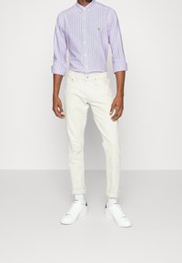 Chemise boutonnée rayée violet clair, pantalon blanc ajusté et baskets blanches avec des accents noirs. La chemise a un petit logo sur la poitrine.