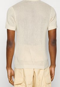 Beżowy krótki sweter z dzianiny z krótkim rękawem, charakteryzujący się teksturowanym wzorem ażurowym. Projekt zawiera ściągacze na rękawach oraz prostą linię dołu.