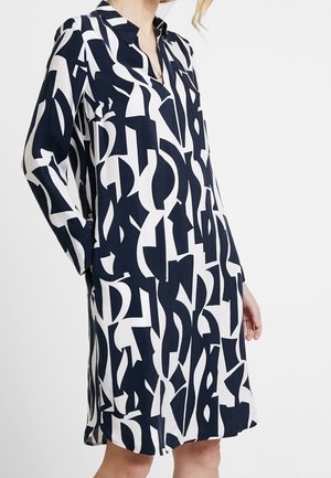 Femme portant une robe à manches longues, longueur genou, avec un motif géométrique abstrait noir et blanc audacieux, debout devant un fond uni.