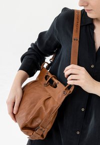Bruine leren tas met een slonzige vorm, verstelbare schouderriem en metalen accenten. De tas heeft een omslag bovenkant met een trekkoordsluiting.