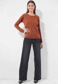 Frau lächelt, trägt einen rostorangefarbenen Langarmpullover, schwarze weit geschnittene Hosen und schwarze spitz zulaufende Schuhe. Sie steht mit einer Hand an der Hüfte vor einer weißen Wand.