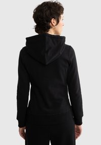 Osoba o krótkich kręconych włosach, ubrana w czarną bluzę z kapturem, widziana od tyłu na jednolitym tle.