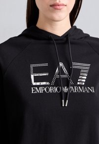 Artimiausias kadras su asmeniu, dėvinčiu juodą gobtuvą su sidabro spalvos „EA7 Emporio Armani“ logotipu krūtinėje ir juodais raišteliais su metaliniais galiukais.