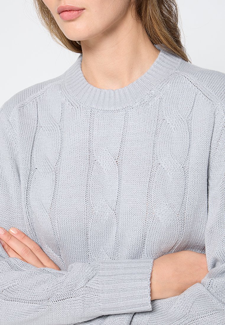 Jersey de punto de cable gris claro con cuello redondo, patrón texturizado y puños acanalados, fabricado en una tela suave. Las mangas son ligeramente holgadas.