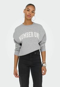 Mujer joven con pendientes de aro, vistiendo un sudadera gris que dice "NÚMERO UNO" y jeans negros de cintura alta, apoyándose contra un fondo neutro.